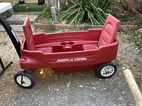 Side view of red Radio Flyer plastic wagon showing fold-up seats and four wheels, outdoors on dirt ground.