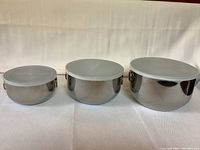 Front view of three stainless steel nesting bowls with lids on a white fabric backdrop, showing varying sizes from small to large.