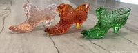 Photo showing all three Fenton cat head glass slippers in pink, orange amber, and green colors on a marble surface.