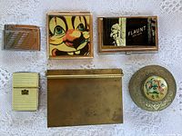 Six items arranged on lace fabric: four lighters, two cigarette cases, one round Persian brass compact on the right.