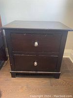 Front view of one dark wood veneer bedside table showing two drawers each with chrome knobs, placed on wooden floor against wall.