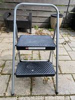 Front view of the collapsible stepping stool showing two black plastic steps with textured ridges and a gray metal frame with a handle.