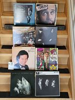 Eight vinyl record albums arranged on stairs showing front covers including George Michael, George Carlin, Julio Iglesias, Stanley Baxter, Genesis, and two unknown albums.