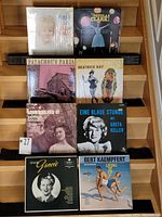 Photo showing seven vintage vinyl records arranged on wooden steps, each in its own jacket or sleeve, covering various artists and classic music themes.