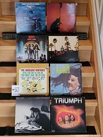 Eight vinyl records displayed on wooden steps, showing front covers. Includes West Bruce & Laing, The Beatles, Kiss - Love Gun, The Ventures, The Fabulous Ventures, Billy Joel - The Stranger, and Triumph.
