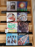 Photo showing seven vinyl record covers arranged on a wooden stair surface. Identified key albums include Ricky Nelson, The Moody Blues, Hugh Masekela, Baron, Three Dog Night.