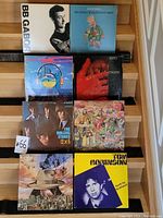 Front view of 8 vinyl records arranged on wood steps showing front cover artwork of BB Gabor, April Wine, Astro Fingers, The Rolling Stones, Iron Butterfly, Weather Report, Tom Robinson.