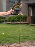 Photo showing the full length of the antique cast iron outdoor bell being held by a person against an outdoor background with green grass and a house in the distance.