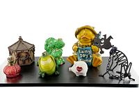 Full view of six kitschy décor items including figurines, piggy banks, and candle holder on a black surface.