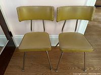 Pair of vintage MCM chairs with tubular chrome legs and olive green vinyl seats and backrests, displayed side by side on wooden floor against pale wall