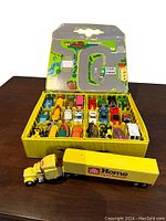 Yellow plastic Matchbox carrying case open showing multiple rows containing 24 vintage die-cast cars and the Home Hardware transport semi truck bank placed in front