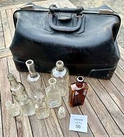 Photo showing large black leather doctor's travel bag with monogram, sits behind a group of 9 vintage bottles of different sizes and shapes on wooden surface.