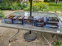 Photo showing multiple stacks of assorted Blu-Ray movie cases arranged on a table outdoors, total around 27 movies in lot.