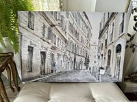 Full front view of the large canvas art depicting an empty European city street with tall buildings and arches in neutral colors, positioned on a white cushioned surface indoors.