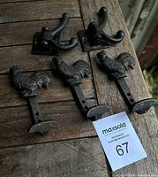 Lot of five cast iron hooks displayed on a wooden surface including three featuring rooster designs and two with double-prong swinging hooks.