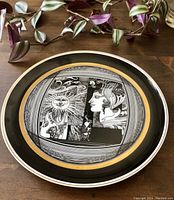Full view of the circular ceramic plate featuring a black and white artistic design with a gold band near the rim, placed on a wooden surface with green and purple plants nearby.