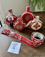 Four folk art embroidered canteen bottles in red, white, and blue on a wooden table with a red embroidered belt laid in front