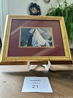 Framed art print showing a stylized mountain peak and cloudy sky in double mat and gilded gold frame, resting on a stand with a plant in the background.