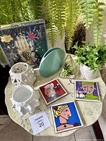Photo showing 9 items on a round table including Christian Lacroix puzzle box, Ursula Dodge art tiles, various planters with faux plants, white geometric candleholder, and robin's egg blue vase.