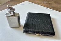 Mini stainless steel flask with screw-top cap alongside black textured cigarette case with gold-tone clasp, displayed on wooden surface.