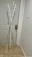 Full view of white IKEA coat rack standing near door, showing tripod three-leg base and multiple hooks.