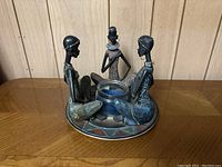 Front angle showing three seated female figurines with painted details surrounding glass votive holder on patterned round base.