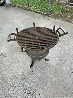 Round metal grill showing weathered rusted surface, primary grilling grate with two wooden handles on sides, on concrete surface outdoors.