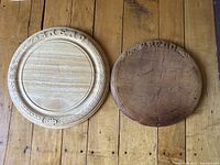 Two round wooden bread boards on wood floor; left board is light-colored with carvings, right board is darker and plain with marks.