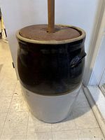 Side view of the two-tone stoneware crock butter churner with wooden lid and long handle.