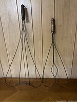 Two carpet beaters with wooden handles and wire frames, standing upright against a wood-paneled wall