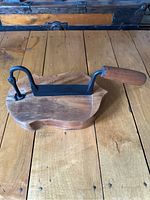 Angled side view of a wooden cutting board shaped like a fruit with a black metal curved blade and wooden handle fixed on one side, placed on a wooden surface.