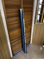 Folded deployable projection screen leaning against wooden doors showing blue metal casing and handle for portability.