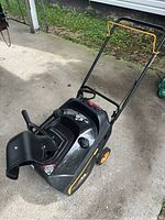 Angled front view of black and yellow single-stage snow blower showing discharge chute, gas cap, and folded handle.