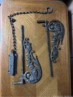 Shows two metal decorative floral scroll gate attachments and one metal chain latch with rectangular latch plate on wood surface