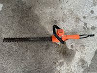 Full view of electric hedge trimmer showing orange and black body with long cutting blade.