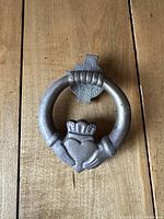 Front view of dark metal door knocker with ring and crowned heart motif on textured wooden surface.