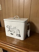 Front view of white metal laundry bin with black vintage text and brush graphic