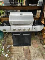 Front view of MasterChef BBQ grill with closed lid showing stainless steel lid, four control knobs on panel, and black cabinet below.