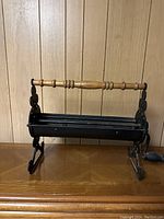 Front view of the cast-iron newspaper roller showing wooden handle and black metal roller drum on a wooden surface against wood-paneled wall.