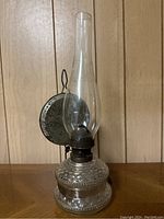 Front view of a vintage oil lantern showing the clear patterned glass base, tall glass chimney, and metal burner with round backing and hook.