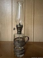 Front side view of vintage oil lantern showing clear glass chimney, metal wick adjuster knob, and metal wrapped glass reservoir with handle