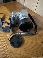 Front view of silver Sony camera with attached lens, neck strap, and detached lens cap on wooden table.