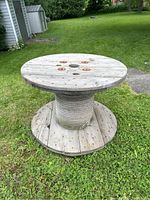 Full view of outdoor wooden spool table standing on grass showing rope wrapped central column and round wooden surfaces top and bottom.