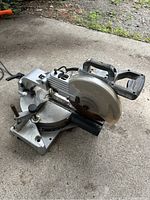 Image of electric miter saw showing side angle with blade cover and handle on outdoor concrete surface.