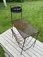 Full view of the outdoor metal side table with perforated surface, built-in ashtray holder on top with ashtray, and magazine rack below, standing on a wooden deck.