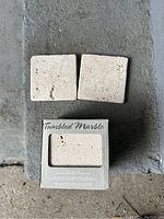 Two individual tumbled marble tiles laid on surface in front of a partially visible box showing the product inside window.