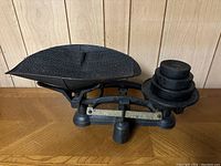 Side view of black cast iron balance scale with large textured pan and weights stacked on the counterbalance side