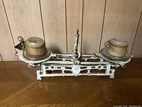 Front view of white cast iron balance scale with two glass plates and pillar candles positioned on each side of the scale.