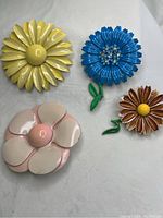 Overhead photo showing all four colorful enamel flower pins arranged on a neutral surface. Yellow, blue with rhinestones, pink and cream, and brown colors visible.