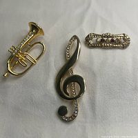 Three gold tone musical brooches displayed on white fabric: a trumpet, a rhinestone treble clef, and an ornate bar pin with red stones.